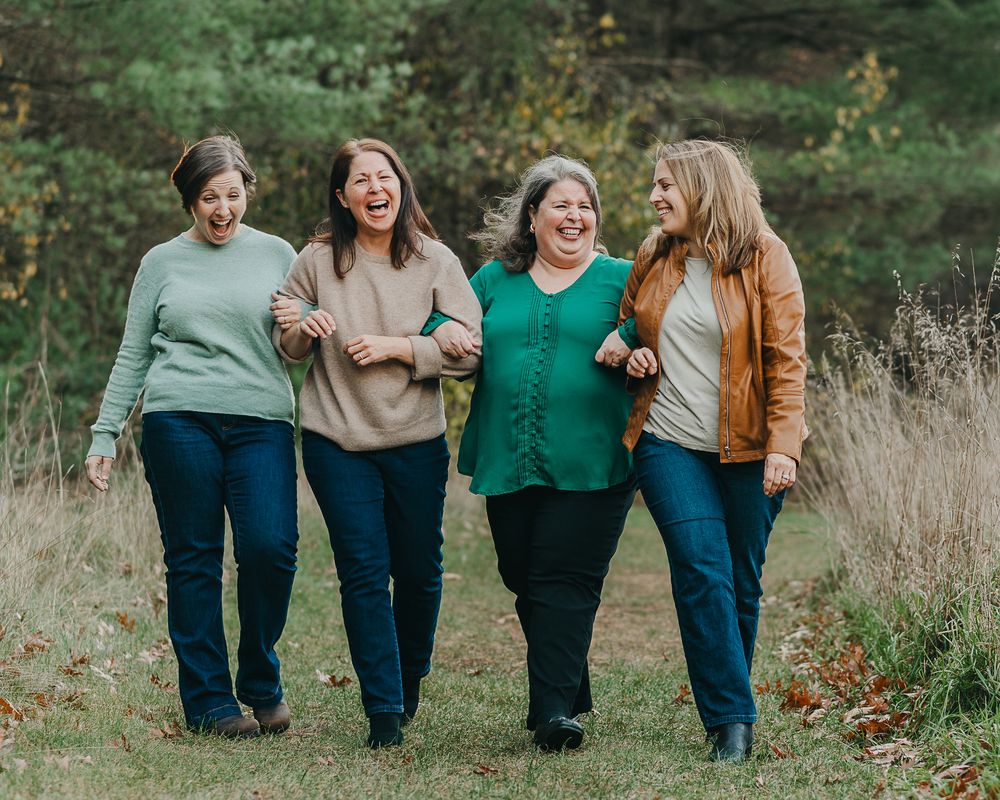 Four grown sisters in casual outfits walks together and laughs in a field among tall autumn grass and foliage.