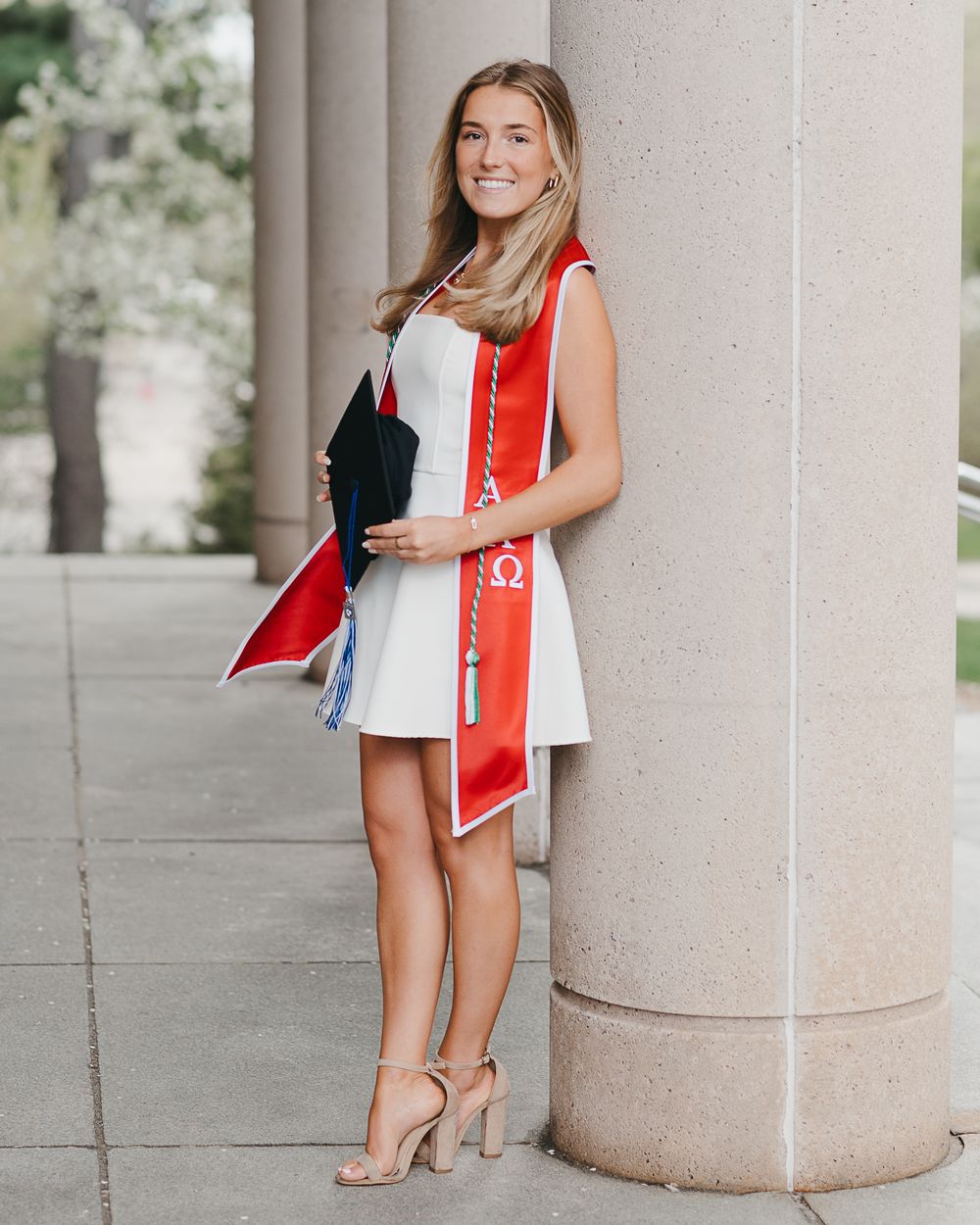 Graduate in white dress with red academic stole stands smiling near stone column in outdoor campus setting.