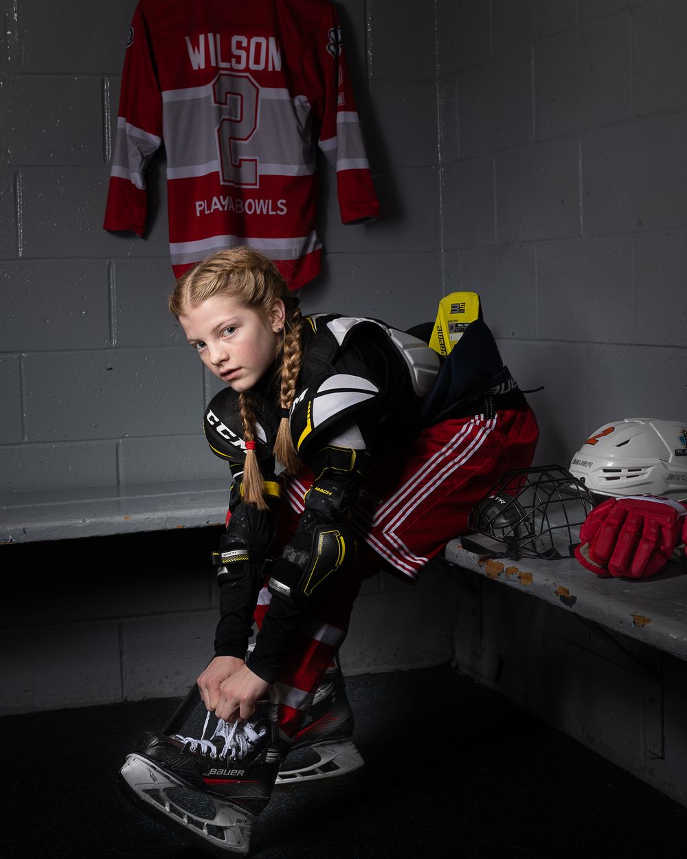 Hockey player in uniform tying skates in locker room with red jersey hanging on wall in background and dramatic lighting