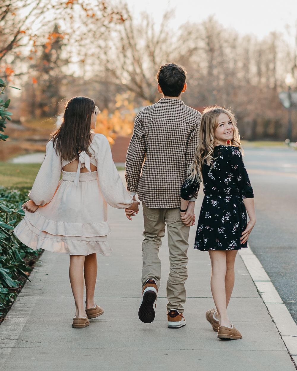 Three siblings holding hands while walking together on a sidewalk in autumn wearing casual dresses and a plaid blazer.