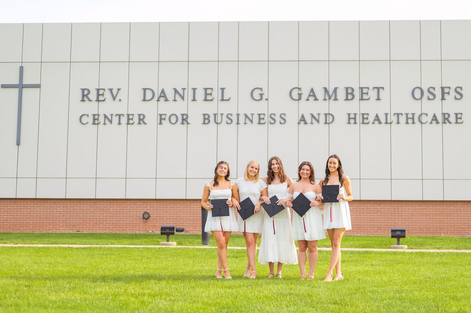 Friends in coordinated white outfits pose together on lawn in front of academic building.