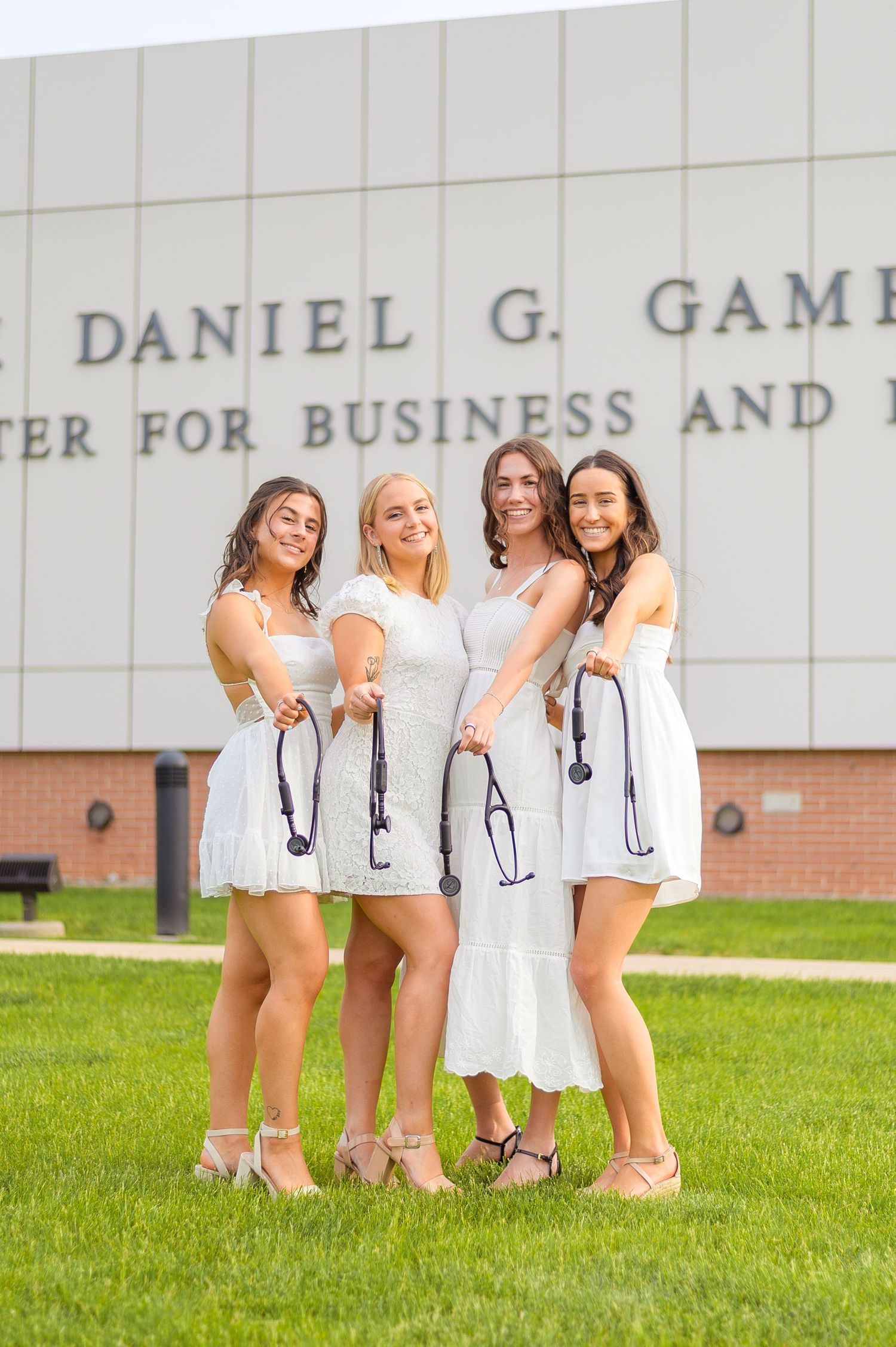 Healthcare graduates holding stethoscopes pose together on grass outside medical center.