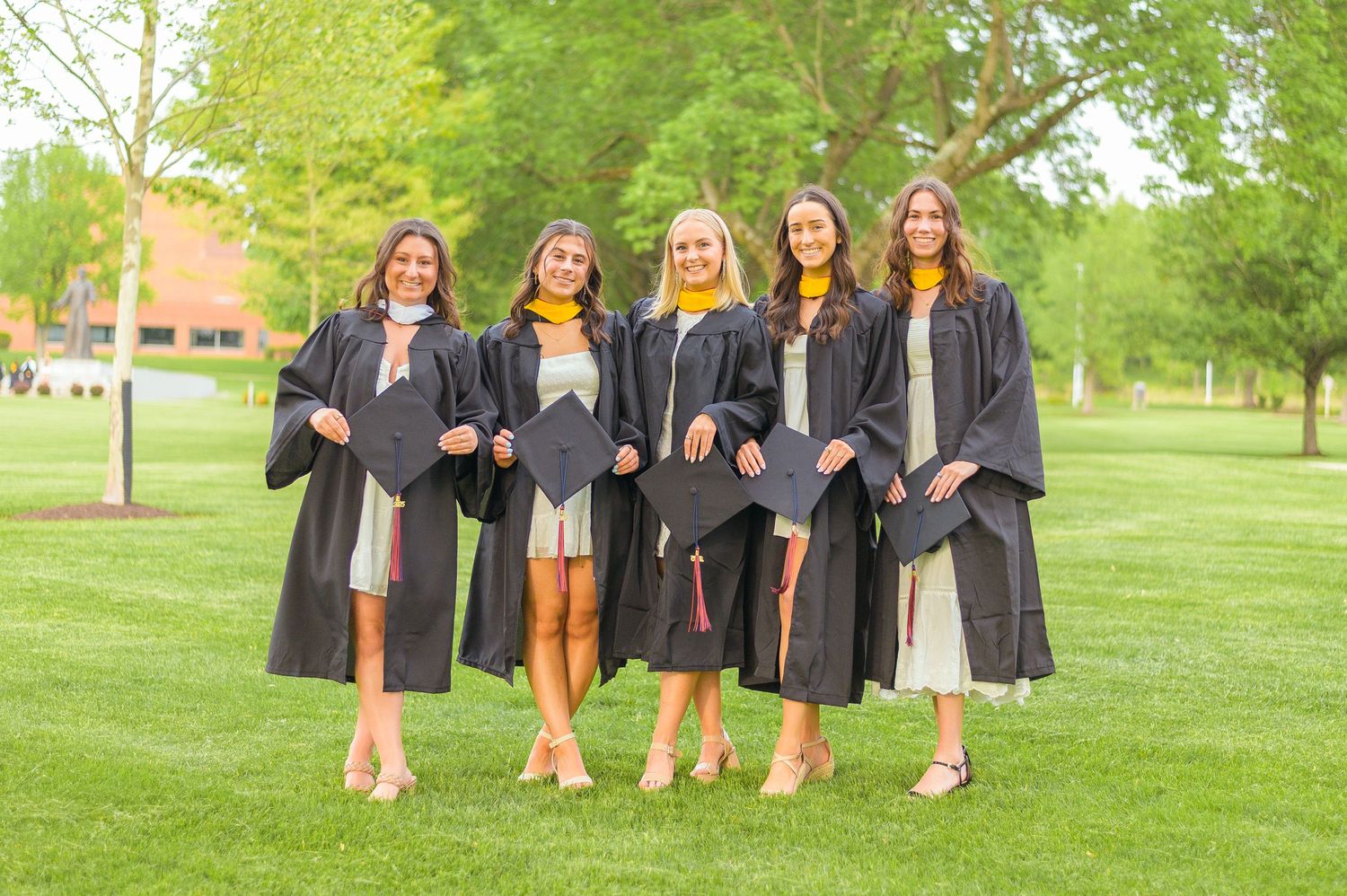 Graduates in black gowns and caps pose on grass with trees and campus buildings in background.