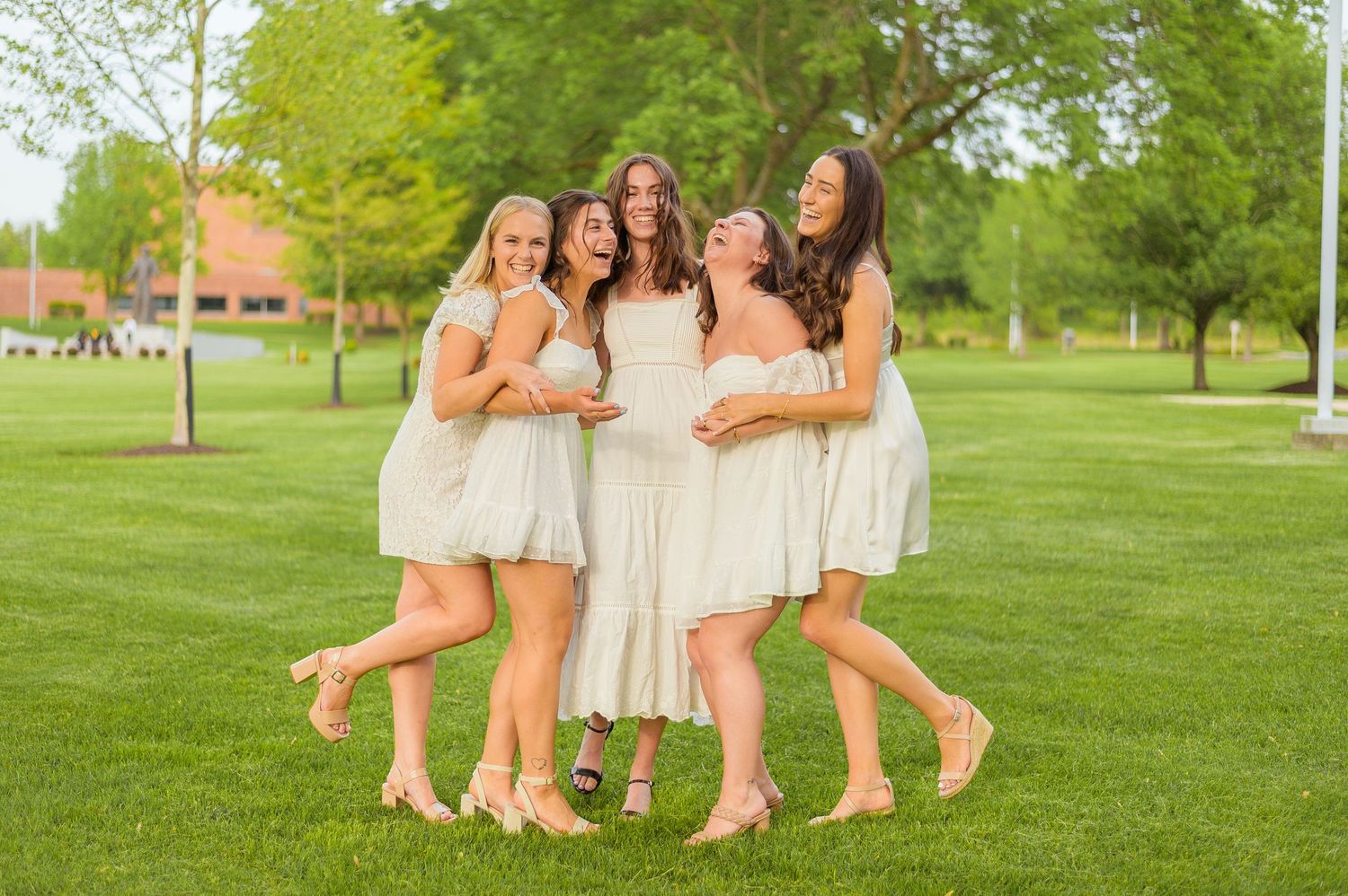 Friends in light summer dresses embrace while celebrating on green campus lawn.