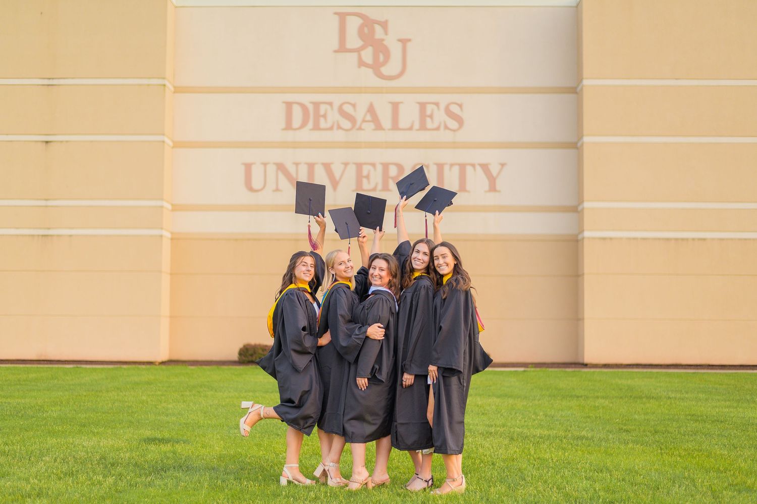 Graduates in black robes pose together in front of DeSales University sign.