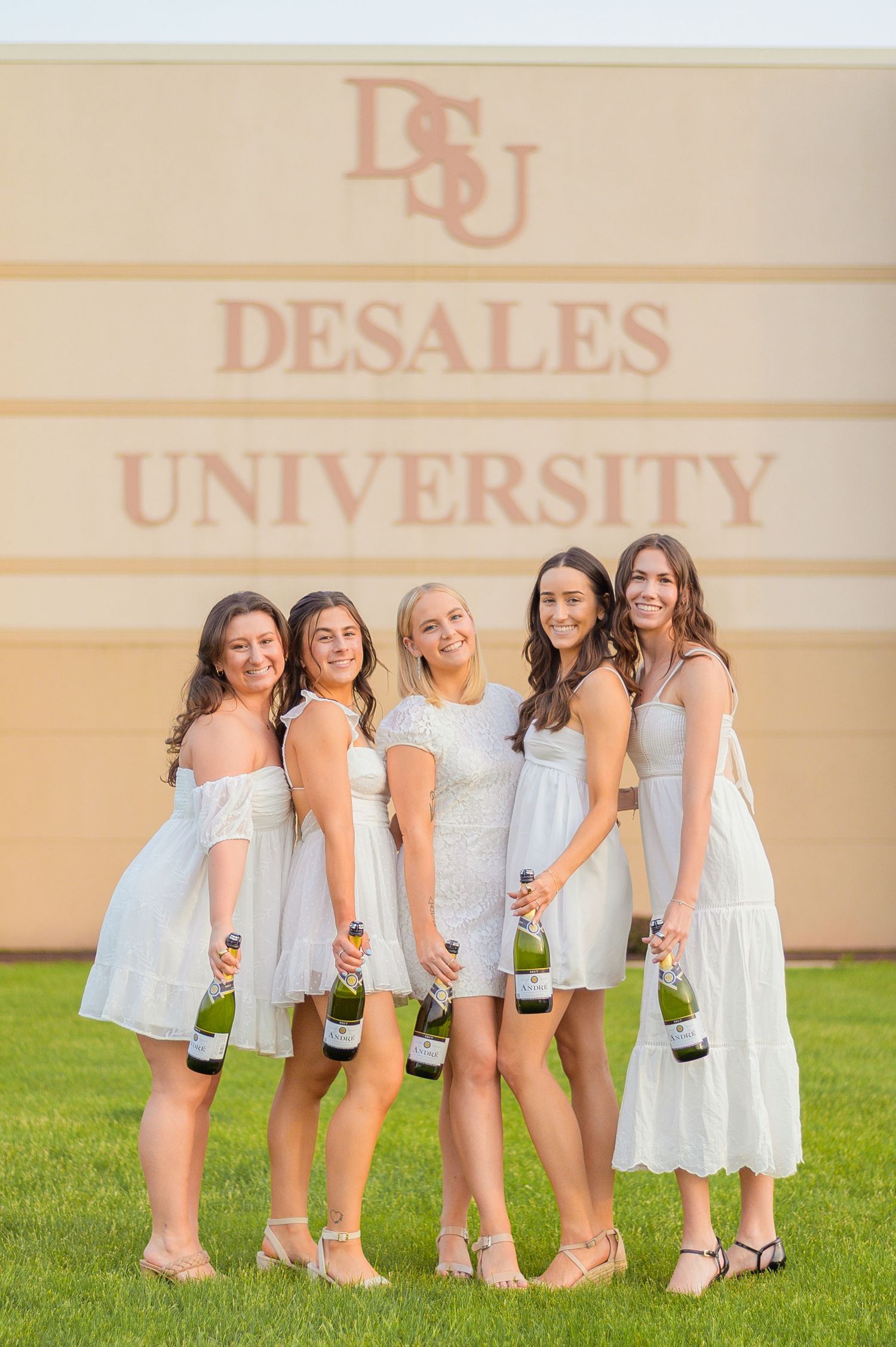 Group celebrating with champagne bottles while wearing white dresses against university backdrop.