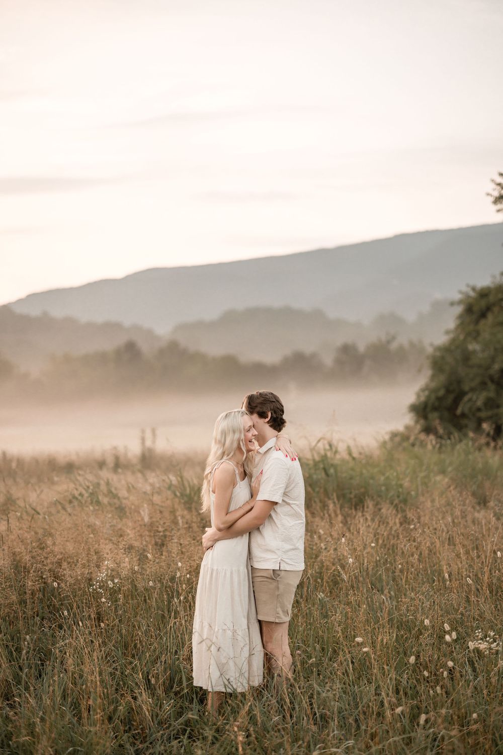 A couple embraces in a misty field of tall grass and wildflowers during golden hour in the mountains.
