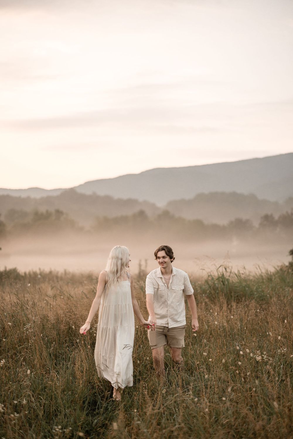 A couple walks hand in hand through a misty mountain meadow at sunset with tall grass and wildflowers.
