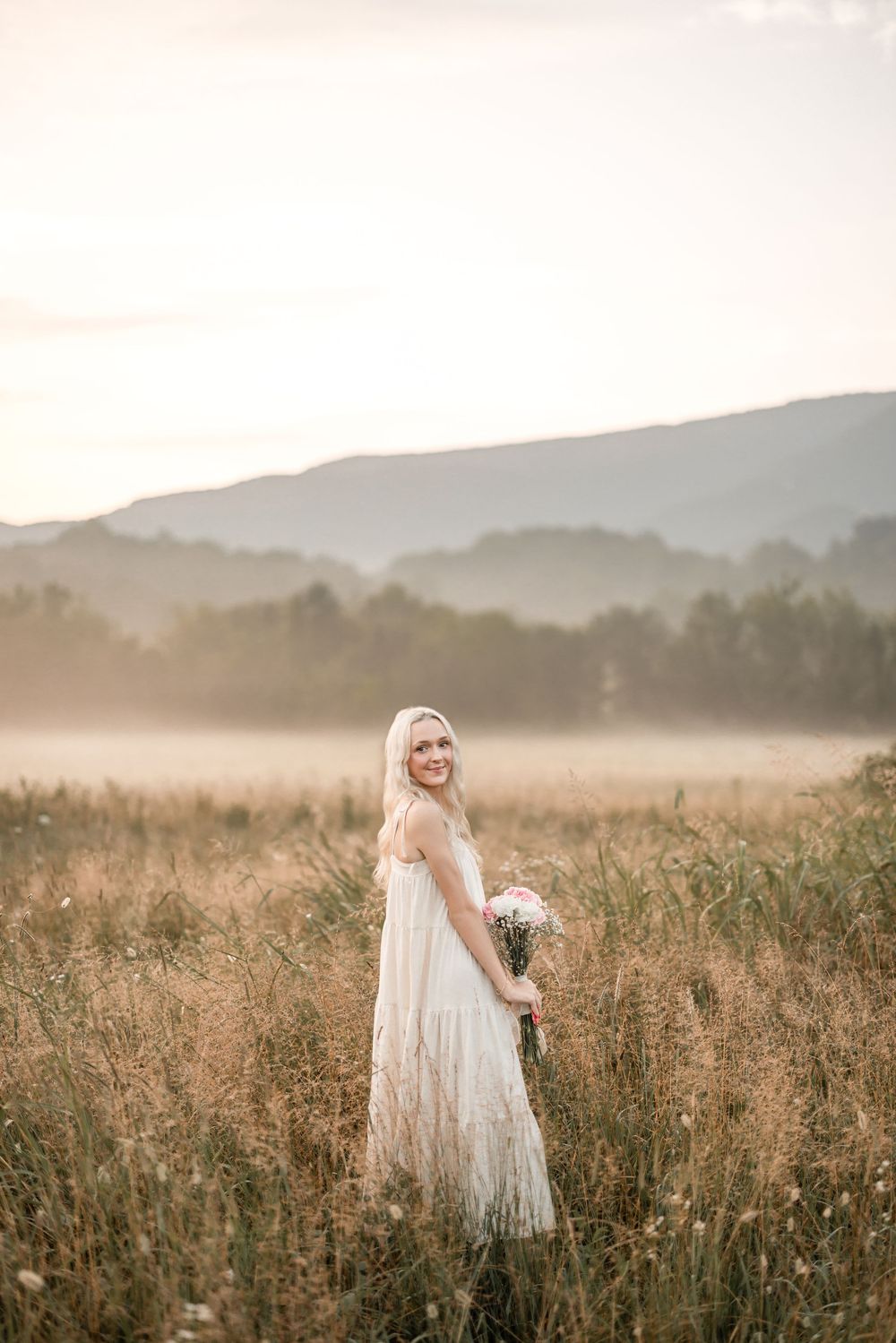 A person in a white lace dress stands in a misty mountain meadow at sunset holding a bouquet of flowers.