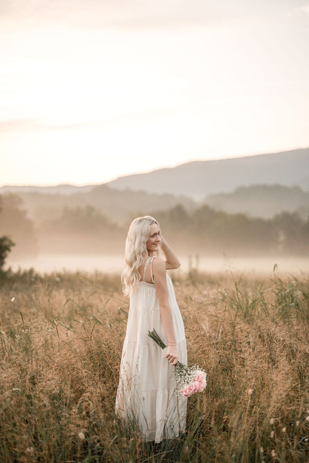 A woman in a flowing white dress stands in a misty field at sunset holding a pink bouquet against mountain silhouettes.