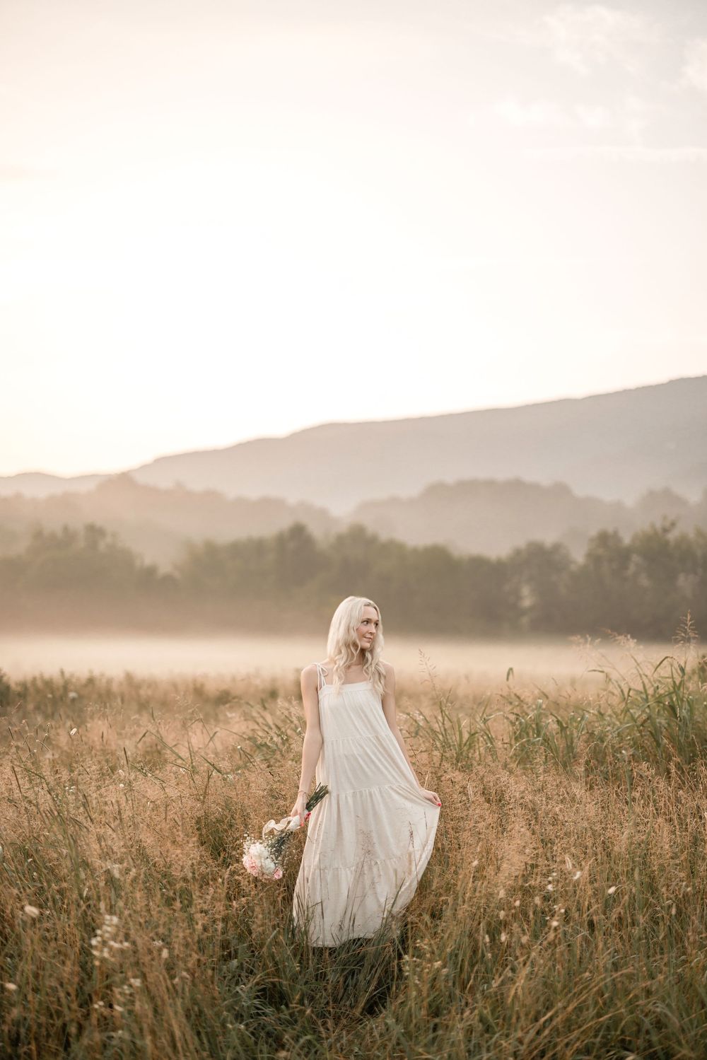 A dreamy sunset portrait in a misty meadow with flowing white dress and mountain backdrop.