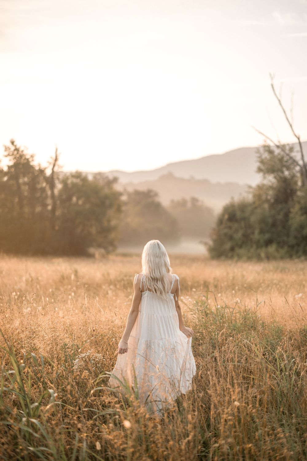 A figure in a white dress walks through a golden field at sunset with mountains in the background.