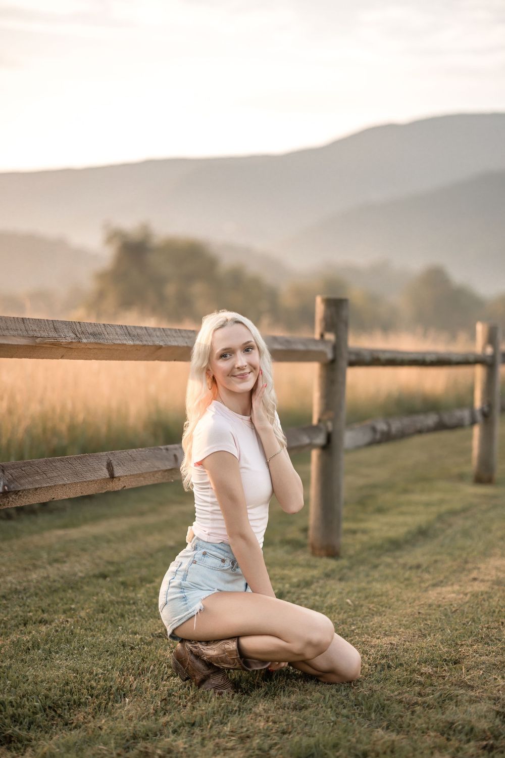 A person poses by a wooden fence in a meadow during golden hour with rolling hills behind.