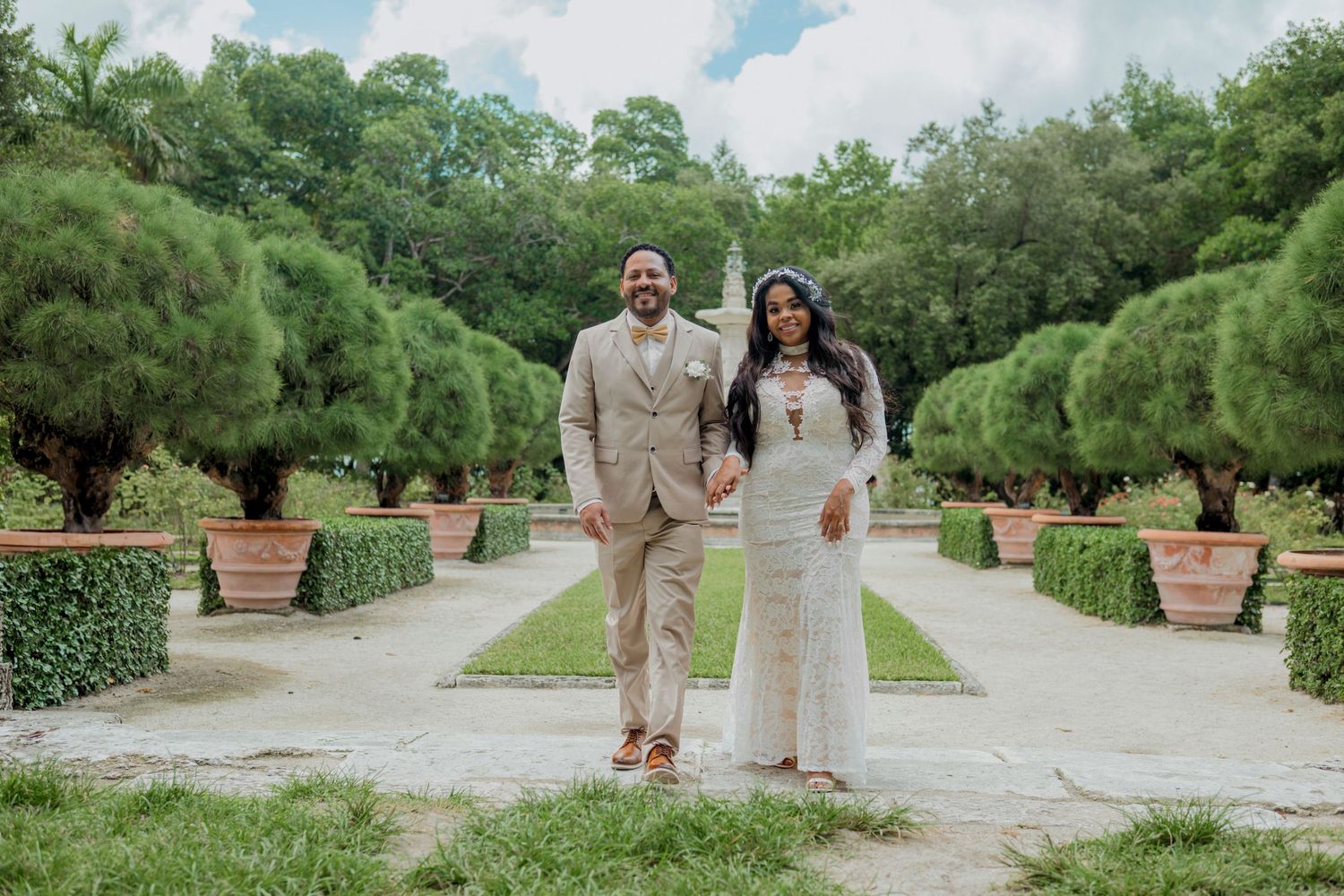 A couple in formal attire stands on a garden path surrounded by manicured hedges and potted plants on a sunny day.