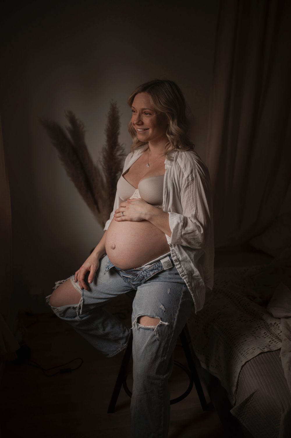 A moody maternity photo of an expectant mother in ripped jeans and white shirt sitting against a dark backdrop with pampas grass.