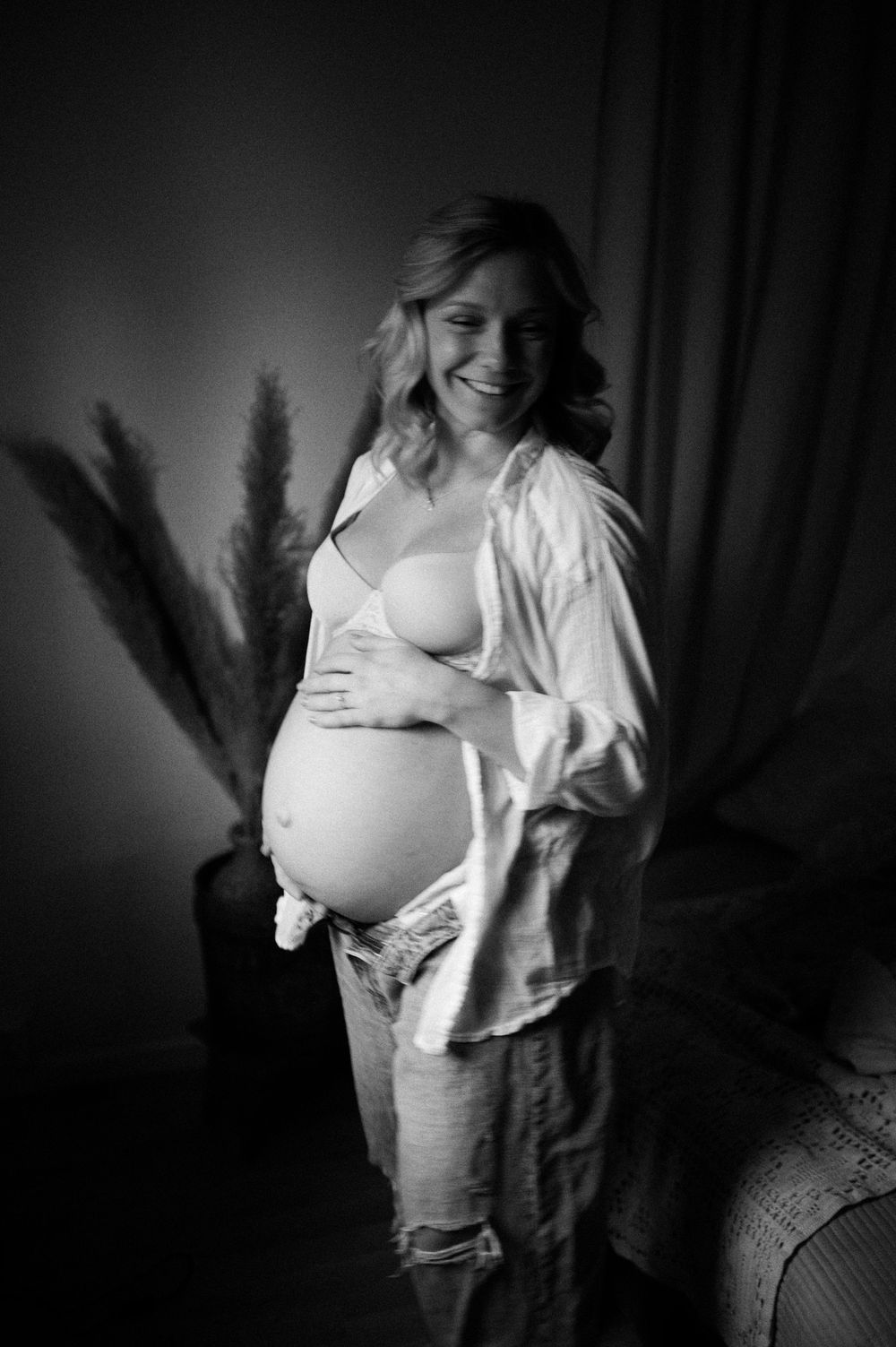 Black and white artistic maternity photo showing expecting mother in white shirt with hands on belly in dark room.