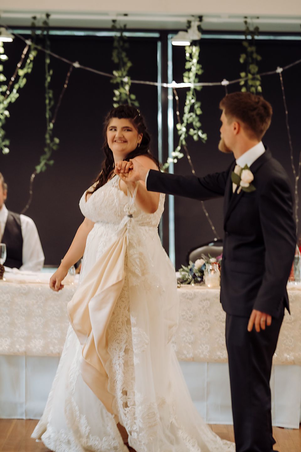 A bride in a flowing white lace wedding dress spins gracefully on the dance floor at an indoor reception.