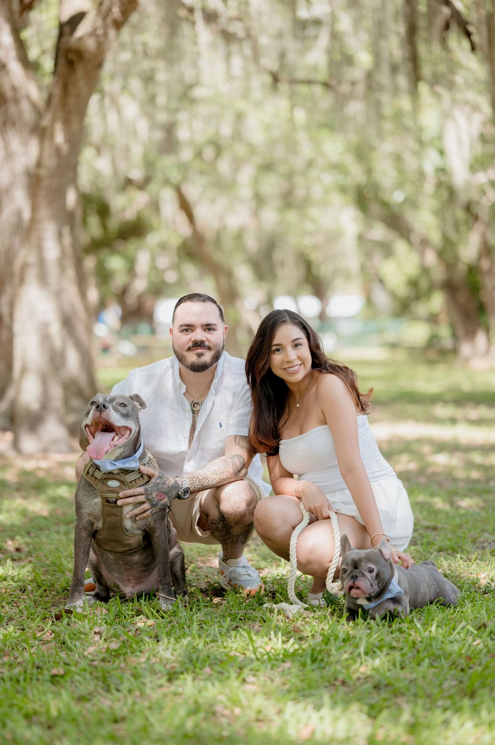 Two people posing outdoors with their pet dogs under large shaded trees in a park setting.