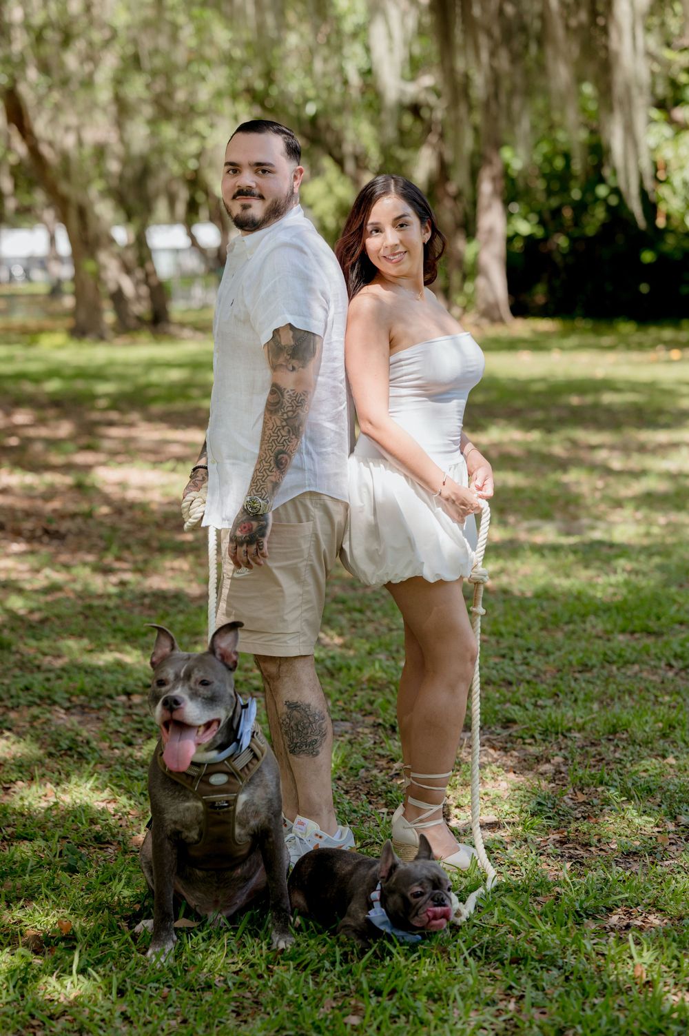 A couple poses back-to-back outdoors in casual attire with their pit bull dog wearing a tactical vest in a park setting.