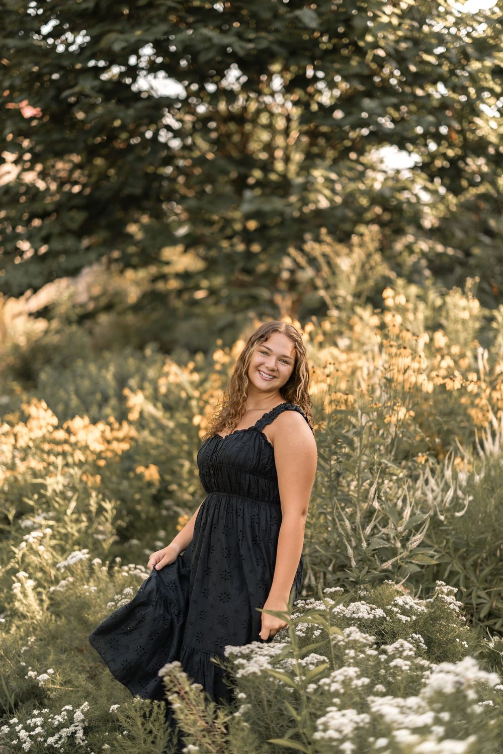 A person in a black dress poses gracefully among wildflowers during golden hour in a natural outdoor setting.