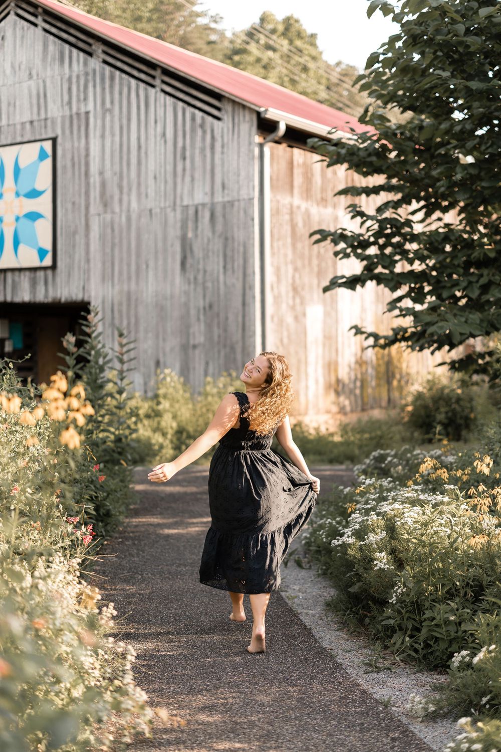 A path with wildflowers leads to a rustic gray barn with a red roof in golden afternoon light.