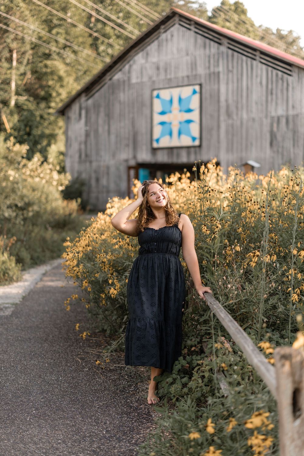 A rustic barn with a blue quilt square pattern stands behind a walkway bordered by golden wildflowers at sunset.