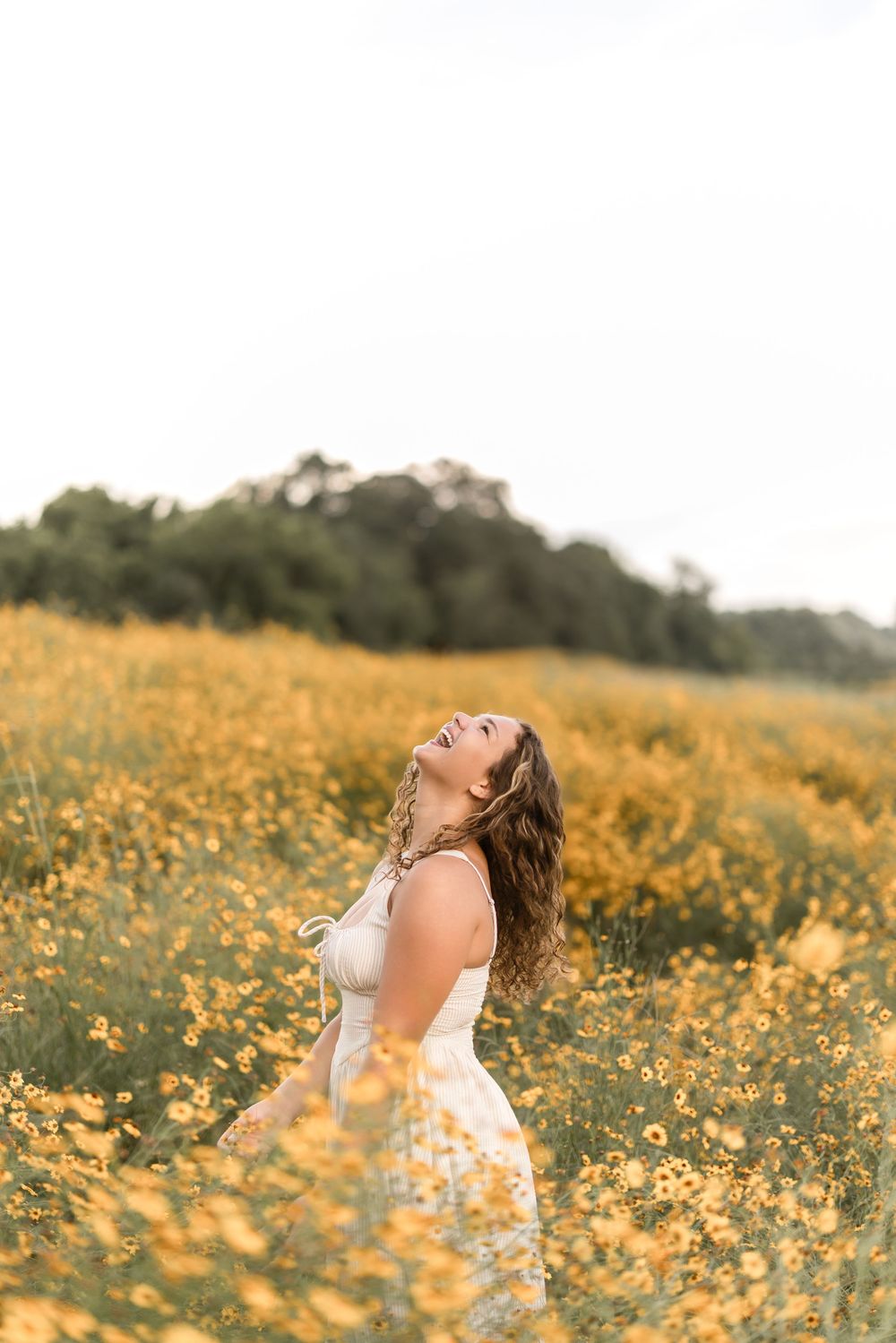 A person in a white dress stands joyfully in a field of yellow wildflowers on a summer day.