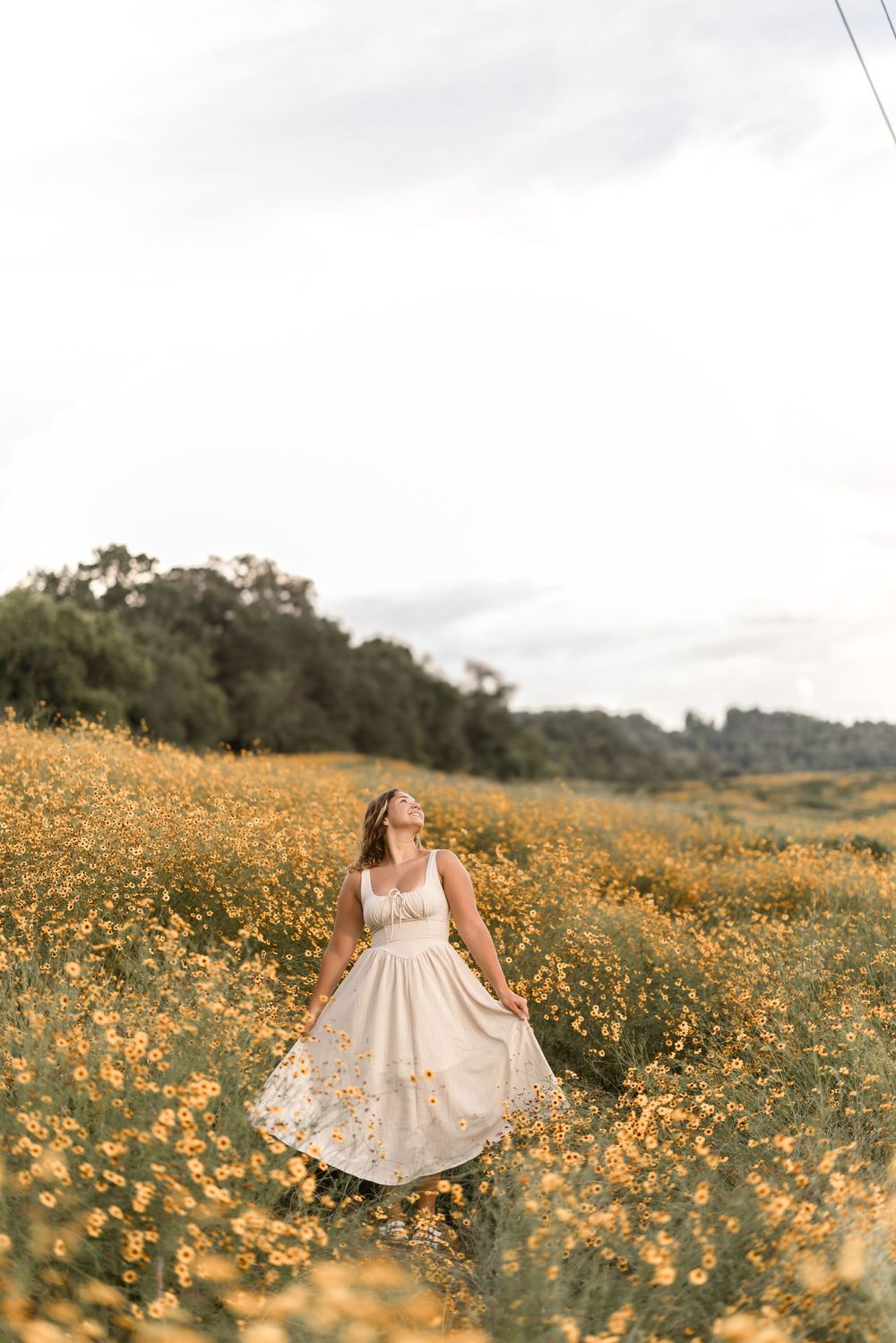A woman in a flowing cream dress twirls among yellow wildflowers in a sunlit meadow with trees in the background.