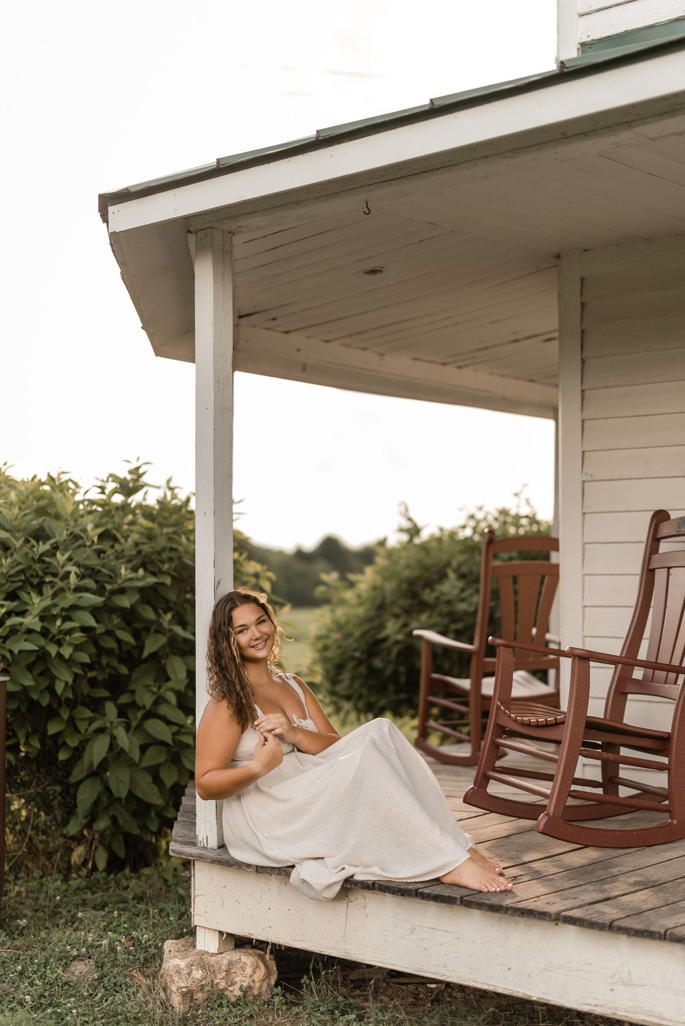 Woman in white dress sits on rustic farmhouse porch with rocking chairs and lush greenery in background.