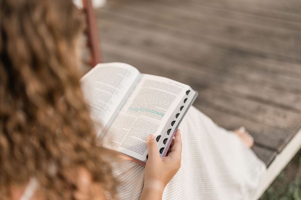 Woman reading a book outdoors on a wooden surface with sunlight filtering through trees in the background.