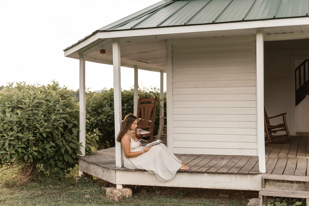 A girl lounges in a white hammock hanging on a farmhouse porch surrounded by greenery and rustic architecture.