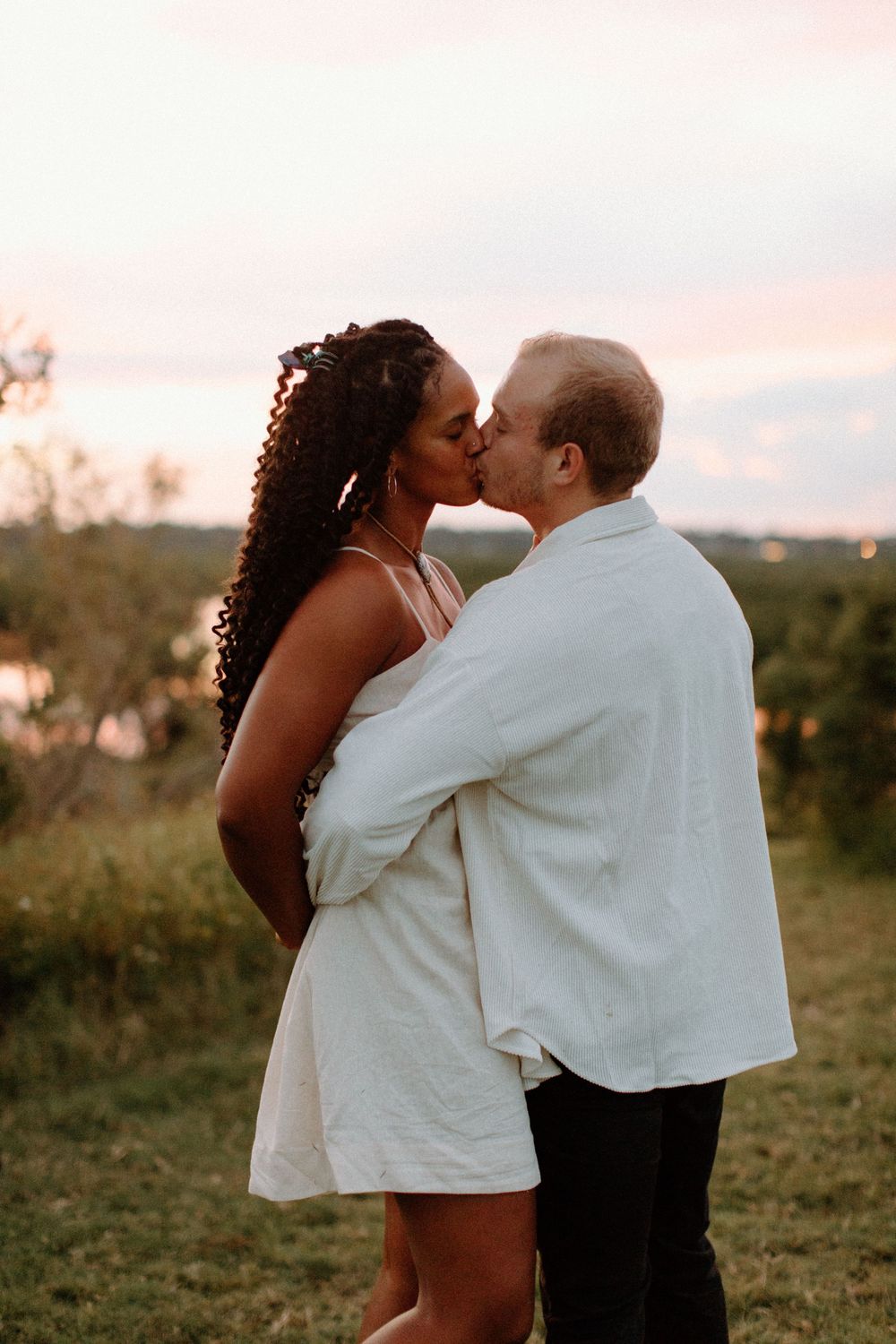 A couple in white clothing embraces while standing in a grassy field at sunset.