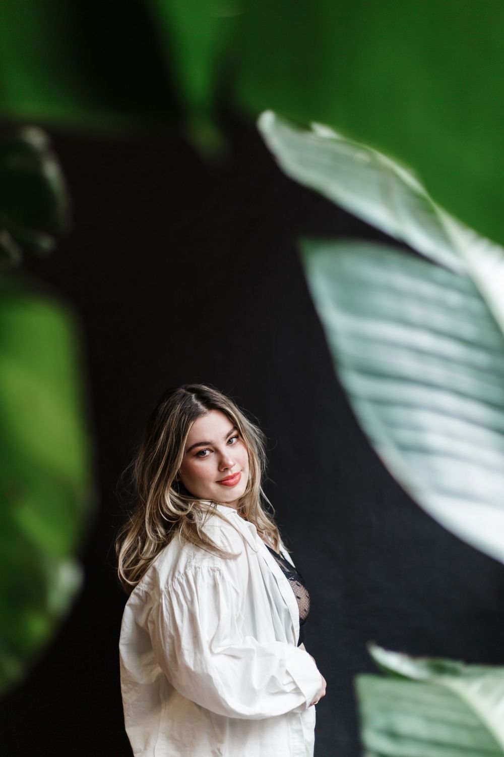 Model in flowy white blouse poses against dark background with large tropical leaves in soft lighting.