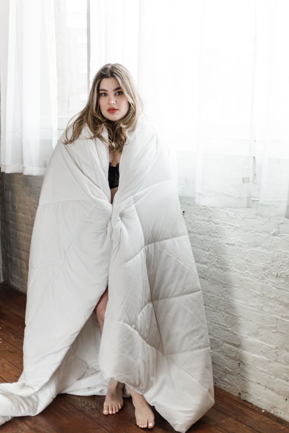 Figure wrapped in plush white comforter sits on wooden floor against brick wall and sheer curtains.