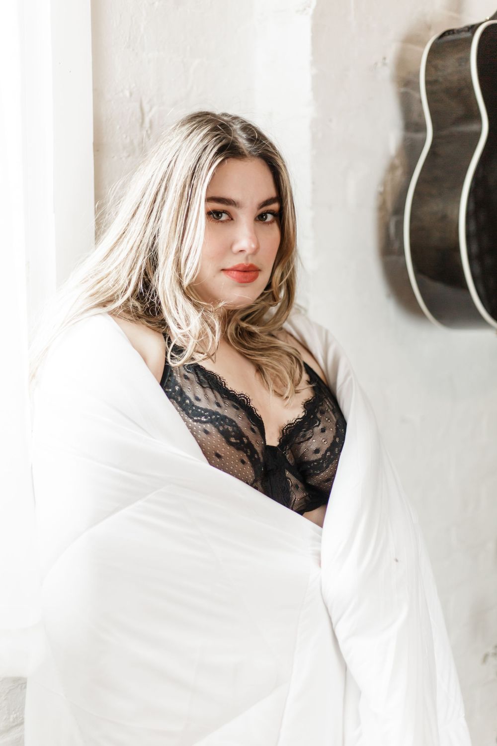 Sensual portrait with black lace lingerie draped in white fabric against light wall with guitar decoration.