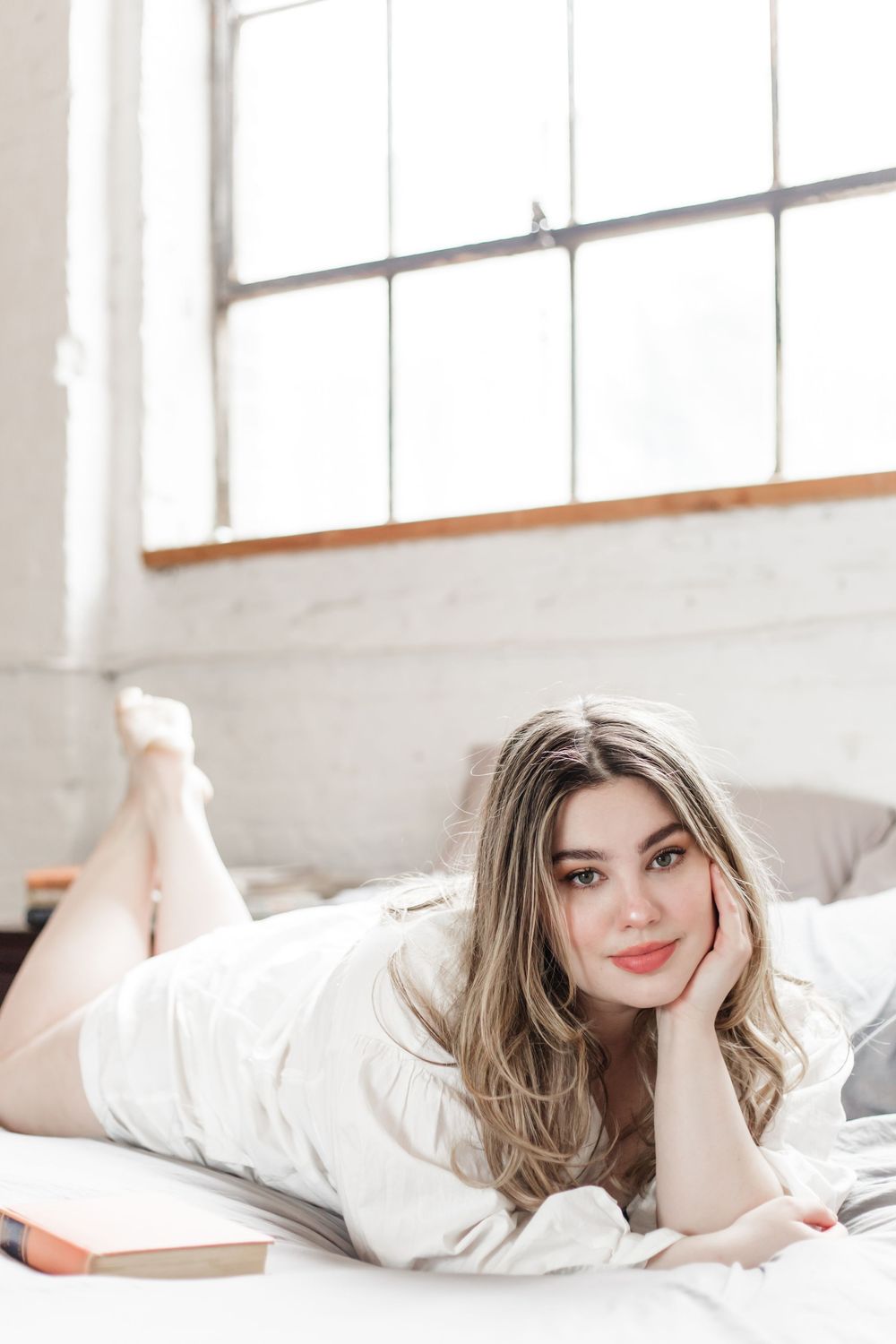 Someone relaxing under white bedding in a bright, sunlit bedroom with windows.