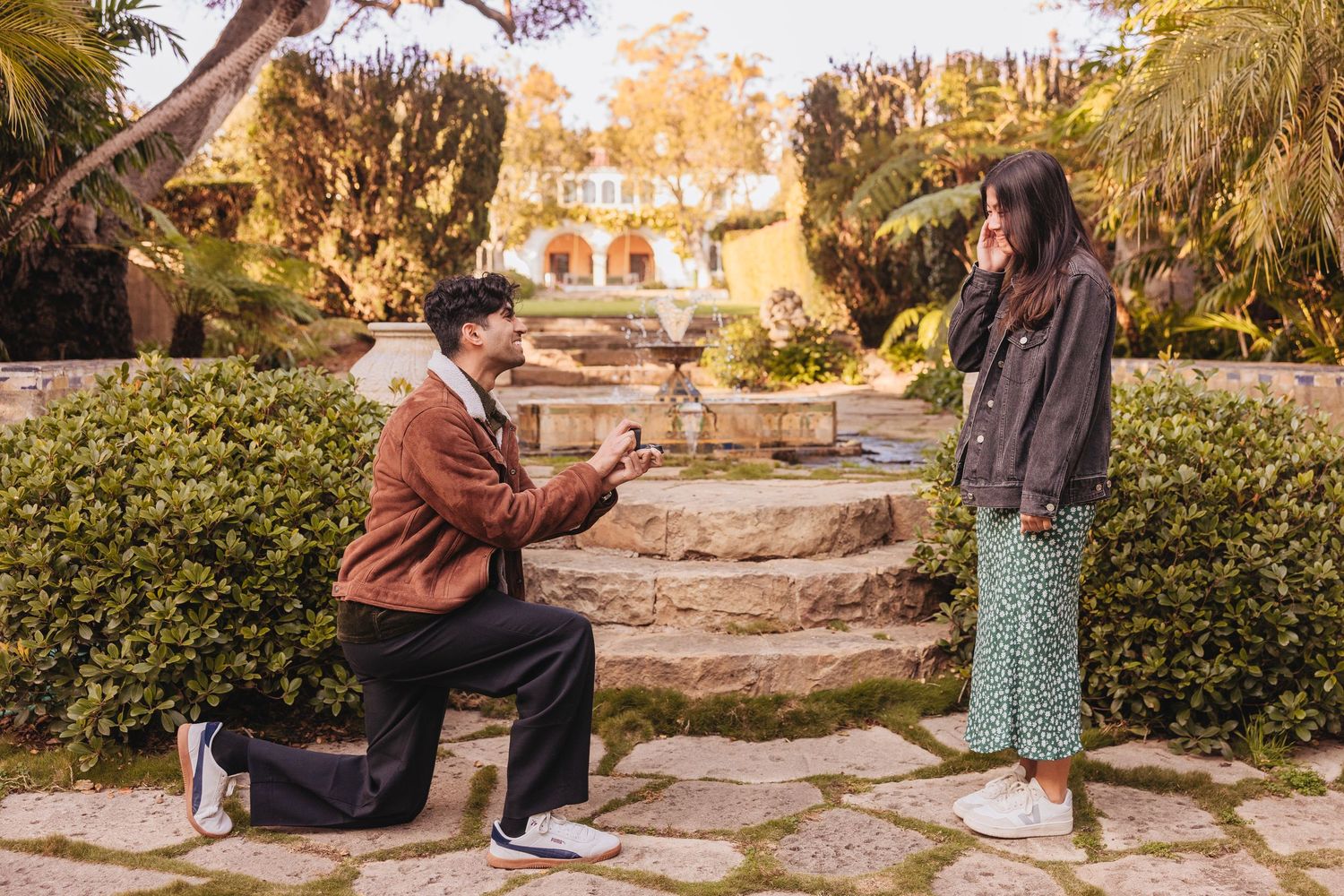 A romantic proposal scene outdoors at sunset with stone steps and lush gardens in the background.