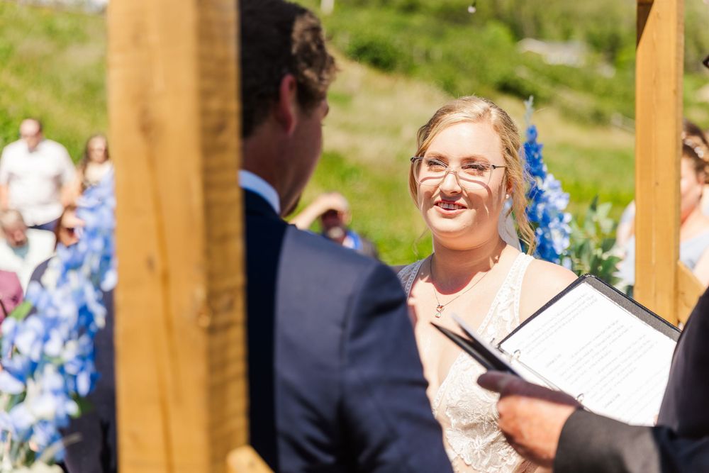A joyful outdoor wedding ceremony takes place under a wooden arch with blue flowers in the background.