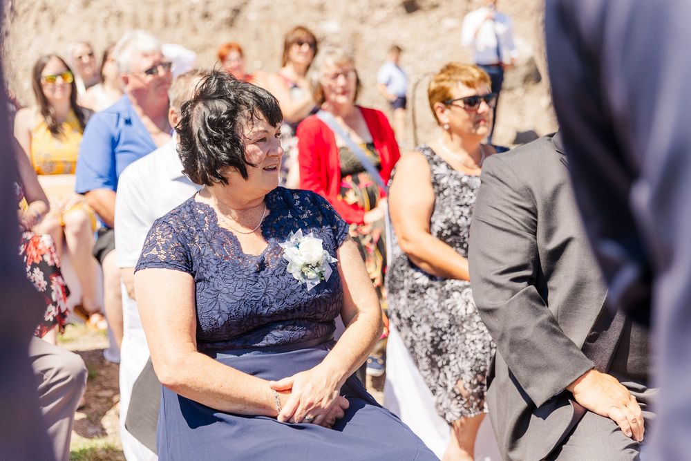 Wedding guests sit in chairs outdoors during a sunny ceremony, wearing formal attire and sunglasses.