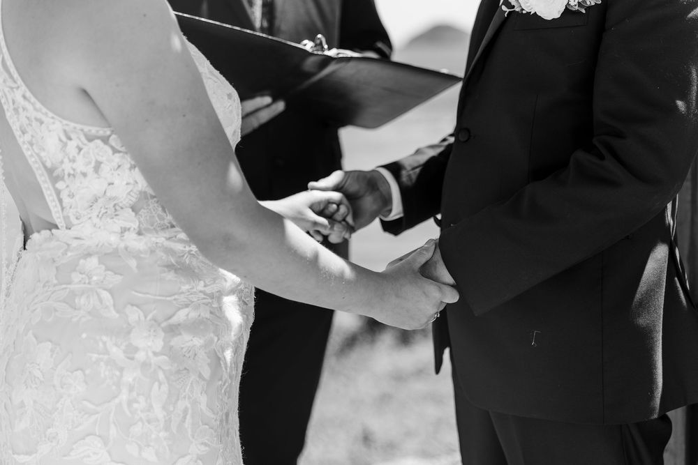A black and white photo showing a couple holding hands at a wedding ceremony, with the focus on their intertwined fingers.