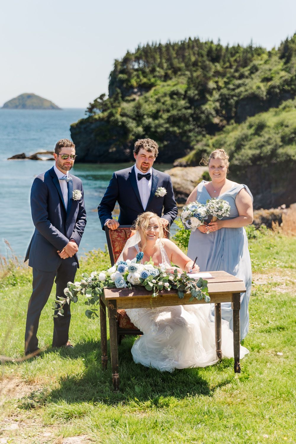 Outdoor wedding ceremony at coastal cliffside location with bridal party gathered around antique wooden table on grassy lawn.