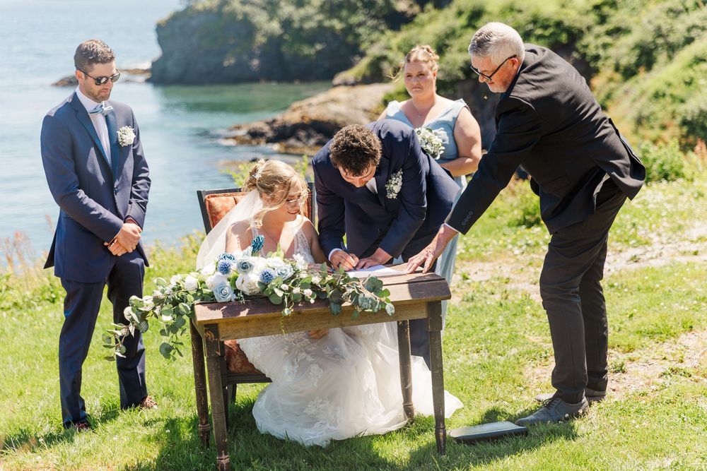 A wedding couple signs their marriage documents in an outdoor ceremony while their celebrant assists.