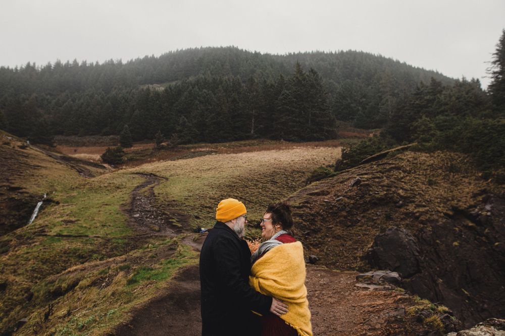 Two people embrace while standing on a cliff overlooking a mountainous landscape.