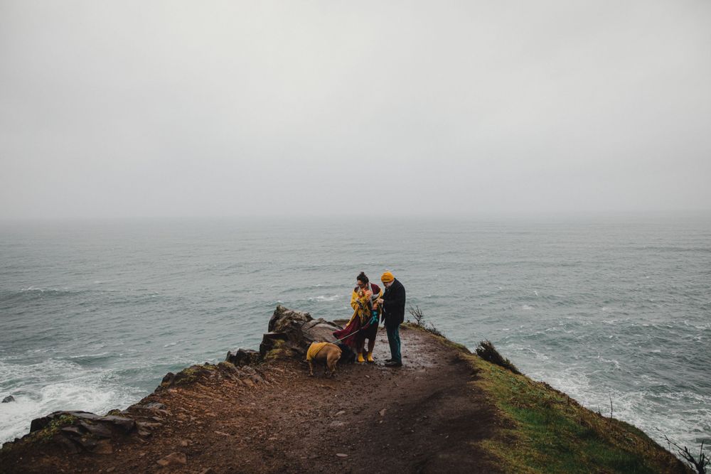 Two figures stand on a dramatic ocean cliff under moody grey skies.