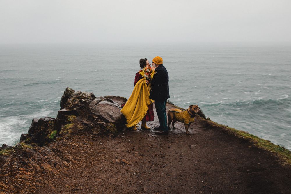 A couple stands with their dog on a dramatic cliff edge overlooking a misty ocean.