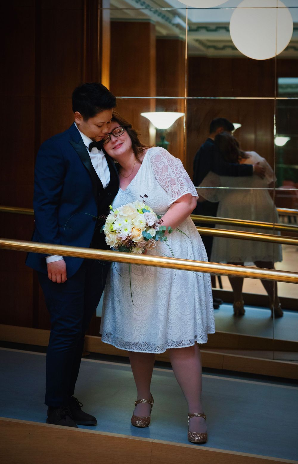 Tender moment between a queer couple in formal wear holding white bouquet in an elegant indoor setting.