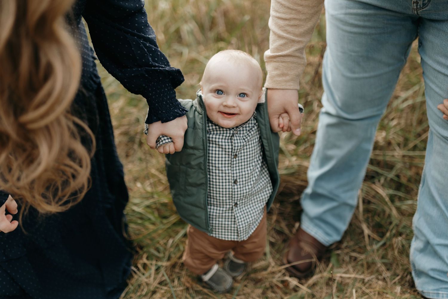 Small child in checkered shirt walking while holding hands with parents in outdoor setting.