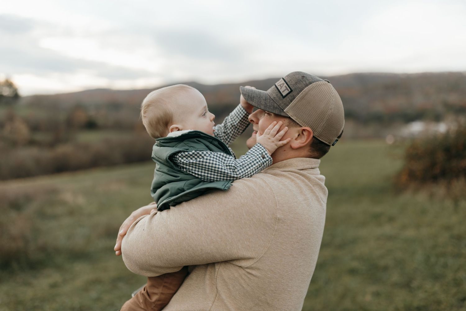 Parent holding and snuggling with baby outdoors against scenic fall landscape.