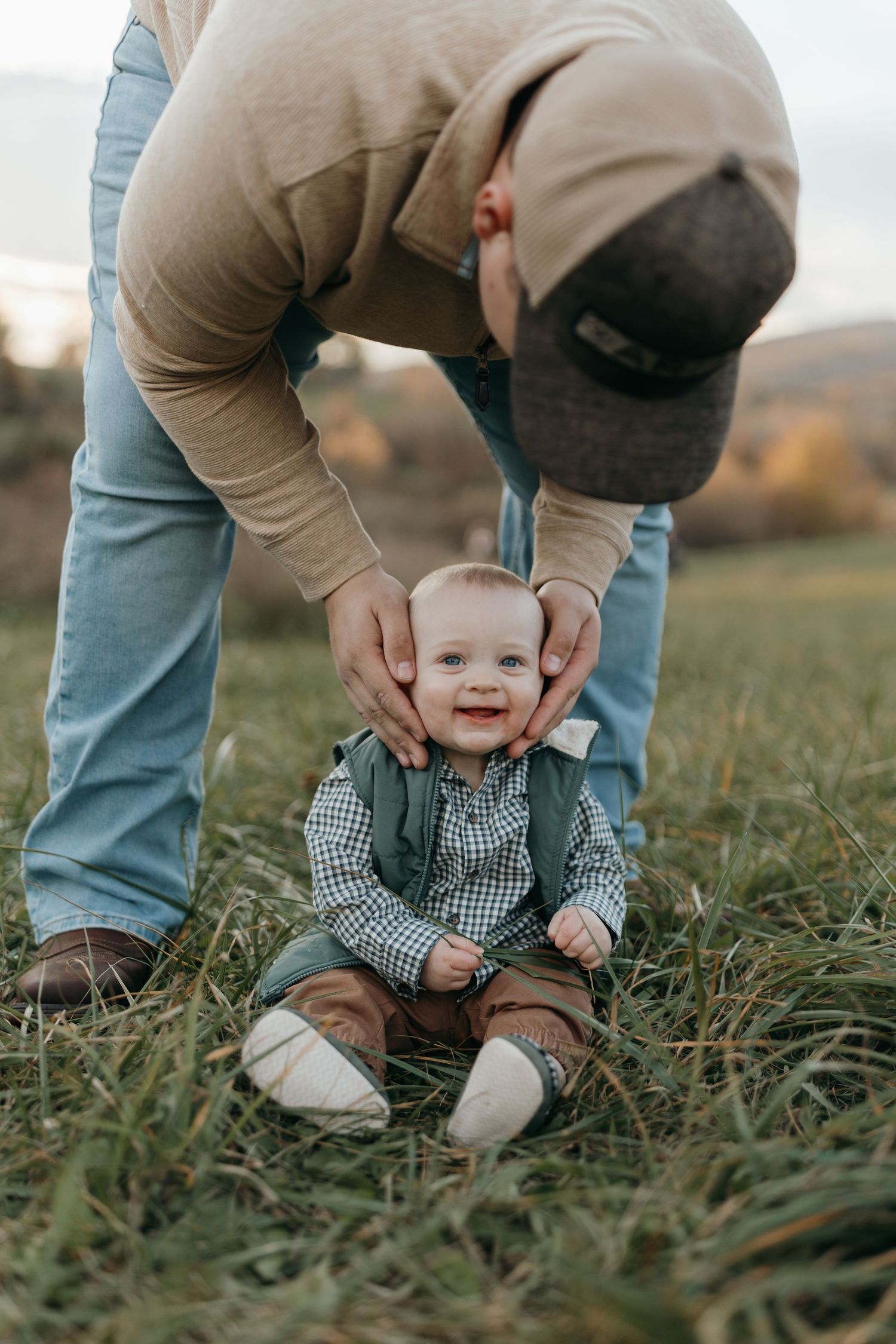 Joyful baby sitting in grass while being held by adult hands during outdoor photoshoot.