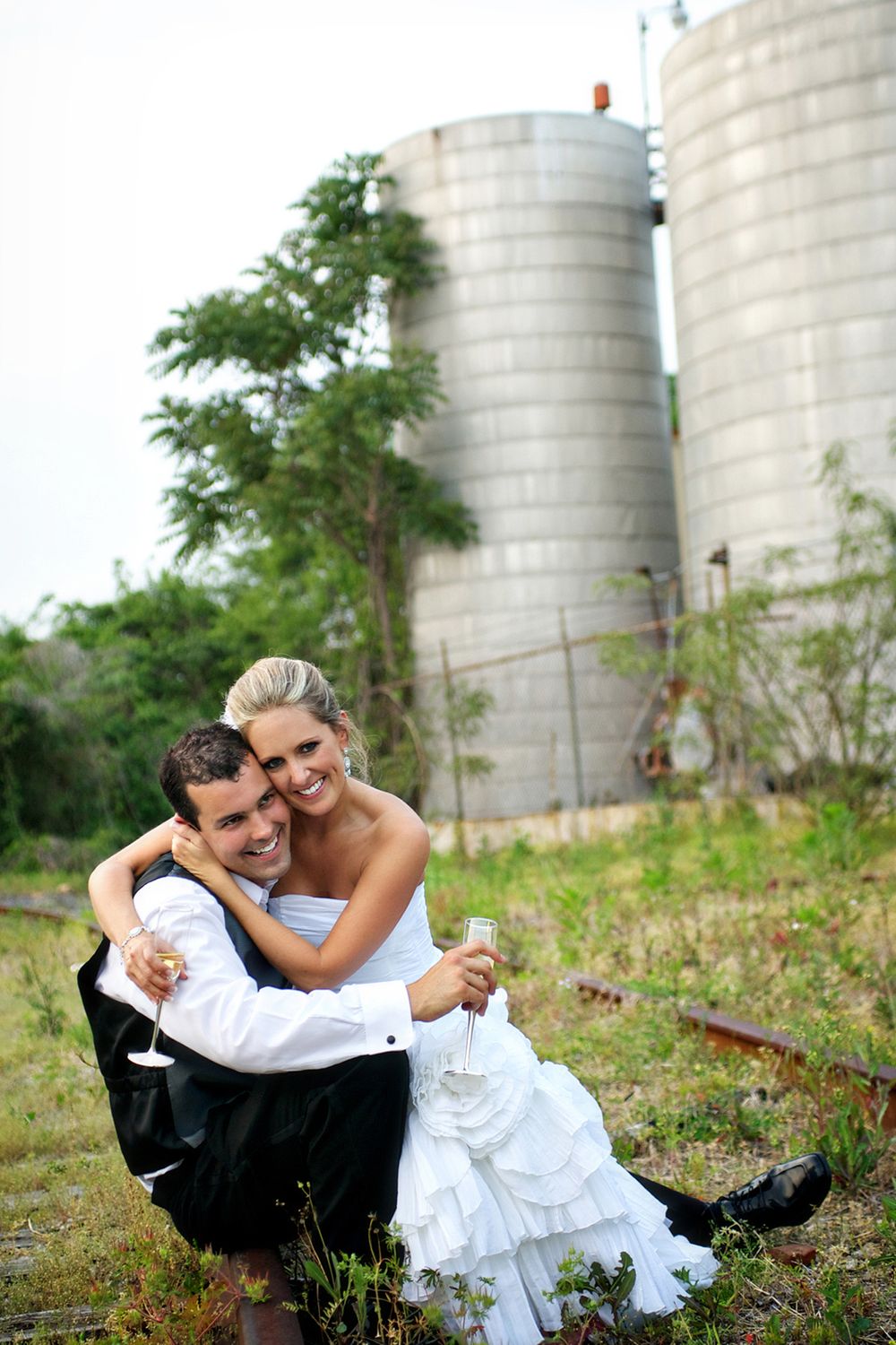 Elegant couple in wedding attire share a romantic moment sitting by industrial silos in a rustic outdoor setting.
